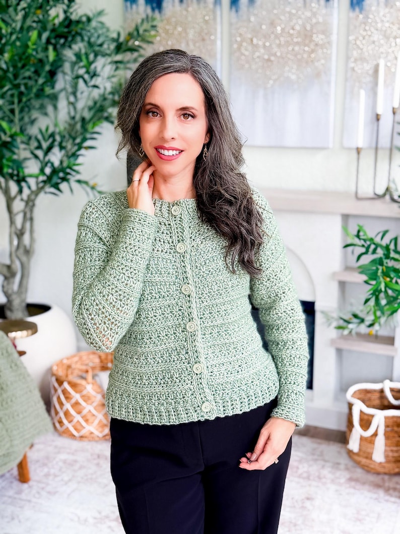 May include: A light green crochet cardigan with a button-up front. The cardigan has a textured stitch pattern and long sleeves. The buttons are a matching light green color. The person is wearing black pants.