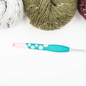 Op de afbeelding: Een haaknaald met een groenblauw handvat met witte stippen en een lichtroze uiteinde, geplaatst onder drie bollen garen in wit, olijfgroen en donkerbordeaux, elk met kleine spikkels van contrasterende kleuren door de gehele vezel heen.