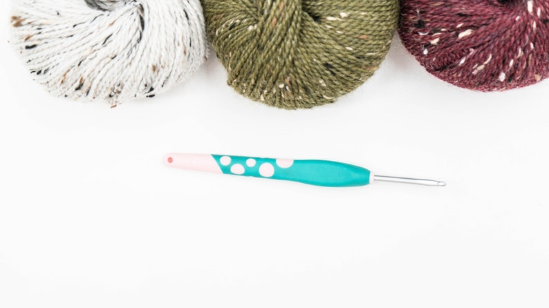 Op de afbeelding: Een haaknaald met een groenblauw handvat met witte stippen en een lichtroze uiteinde, geplaatst onder drie bollen garen in wit, olijfgroen en donkerbordeaux, elk met kleine spikkels van contrasterende kleuren door de gehele vezel heen.