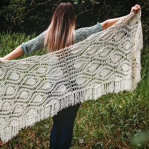 A woman wearing a white shawl decorated with the intricate crochet pineapple stitch, with fringed edges. She stands in an outdoor green space, surrounded by greenery and sunshine.