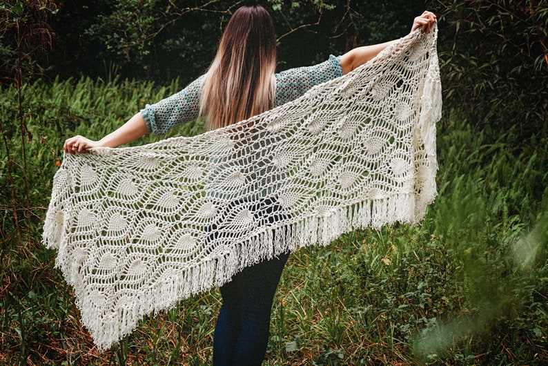 A woman wearing a white shawl decorated with the intricate crochet pineapple stitch, with fringed edges. She stands in an outdoor green space, surrounded by greenery and sunshine.