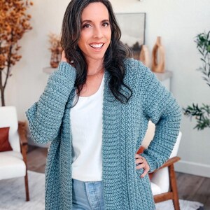 May include: A long, open-front, blue-green crochet cardigan. The cardigan has a textured, ribbed design and long sleeves. The person in the image is wearing a white top and blue jeans. The cardigan is a handmade item.