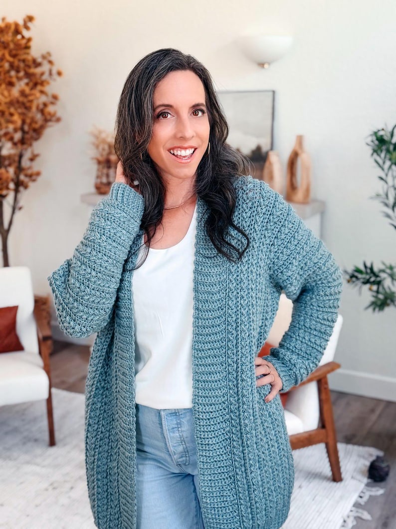 May include: A long, open-front, blue-green crochet cardigan. The cardigan has a textured, ribbed design and long sleeves. The person in the image is wearing a white top and blue jeans. The cardigan is a handmade item.