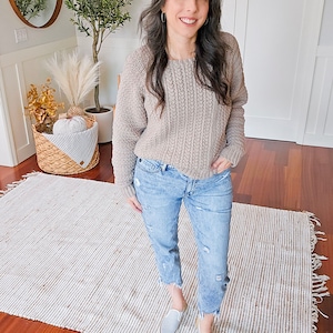 A woman stands on a rug in a room with wooden flooring, wearing a beige knit sweater, blue jeans, and grey slip-on shoes. She complements her look with unique jewellery. In the background, there is a plant and a decorative basket.