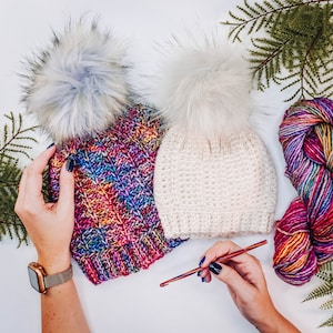 May include: Two crocheted hats with faux fur pom poms. One hat is white and the other is a multi-colored yarn with a rainbow pattern. A crochet hook is on the table.