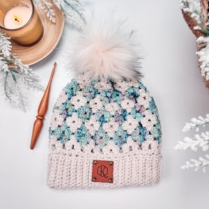 Op de afbeelding: Een gehaakte muts met een crèmekleurige geribbelde boord en een bruin leren label met de letters KR. Het hoofdgedeelte heeft een granny square patroon in crème, blauwgroen en blauw gespikkeld garen, afgewerkt met een pluizige lichtroze pompon.