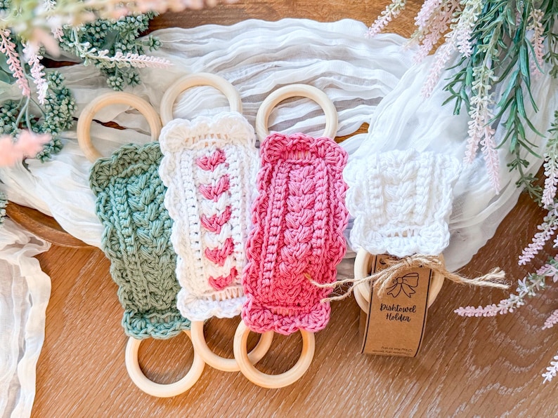 May include: Four handmade crochet dish towel holders in green, white, and pink, each with a wooden ring. The white holder has a tag that reads "Dish towel Holder". The holders are displayed on a white cloth with floral accents.