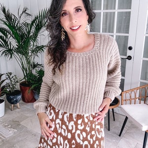 Woman in a beige crochet sweater and animal print skirt, wearing unique jewellery, smiling in a well-lit room with plants and furniture.
