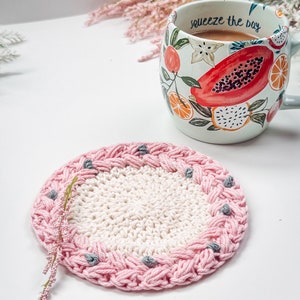 May include: A pink and white crocheted coaster with a gray border and gray beads. The coaster is sitting on a white surface with a mug that says "squeeze the day" in the background.