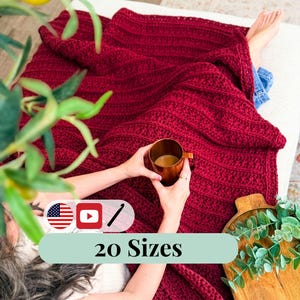 Puede incluir: Una manta de ganchillo roja oscura y texturizada cubre a una persona relajándose sobre una superficie blanca. La persona sostiene una taza de cobre. La imagen incluye el texto "20 Tallas" y un icono de la bandera estadounidense.