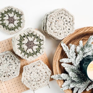 May include: A collection of hexagonal crochet coasters in cream, sage green, and dark brown yarn. Some coasters feature a solid cream color, while others display a multi-colored floral pattern. The coasters are arranged in stacks and individually.