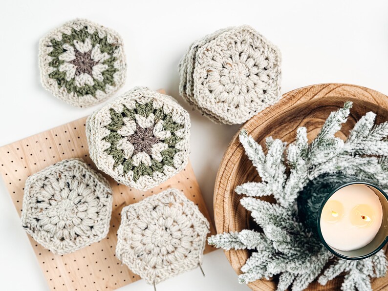 May include: A collection of hexagonal crochet coasters in cream, sage green, and dark brown yarn. Some coasters feature a solid cream color, while others display a multi-colored floral pattern. The coasters are arranged in stacks and individually.