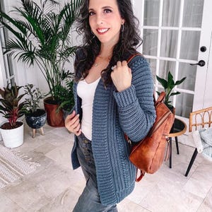 Woman with long hair, wearing a blue crochet cardigan and jeans, holds a brown backpack. She stands in a room adorned with potted plants and a glass door, exuding charm like unique jewelry complementing vintage clothes.