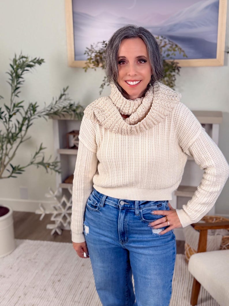 May include: A woman wearing a cream-coloured knit sweater with a cowl neck and blue jeans. The sweater has a textured pattern. The jeans are a light wash with distressed details. The woman is smiling.
