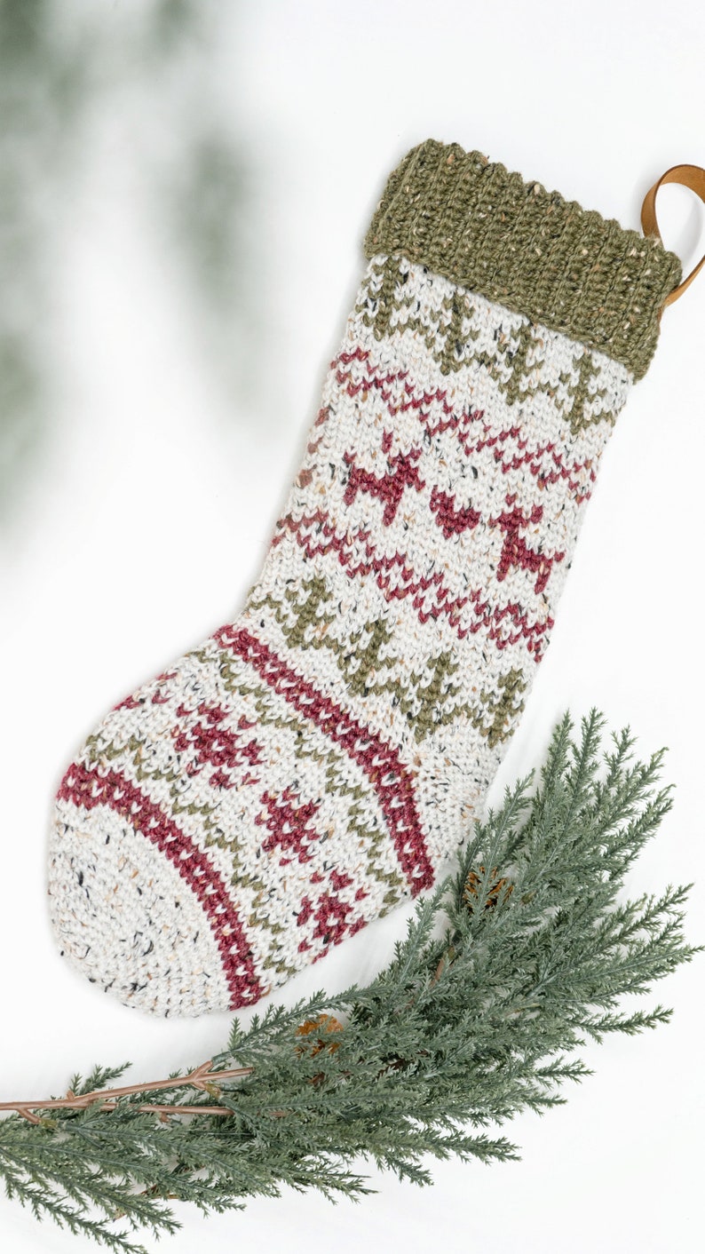Op de afbeelding: Een gebreide kerstsok met een gespikkelde cr&egrave;mekleurige basis en herhalende patronen van donkerrode rendieren en olijfgroene dennenbomen. De bovenste boord is van olijfgroen geribbeld breiwerk en heeft een klein bruin leren lusje om op te hangen.