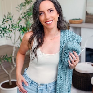 May include: A woman wearing a cream-colored tank top and light blue jeans, holding a teal-colored crocheted shawl and a ball of yarn. A green plant is in the background.