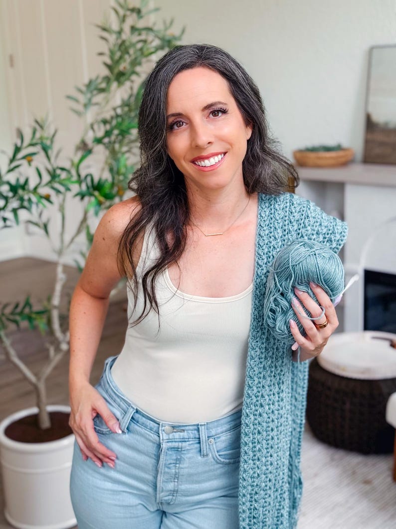 May include: A woman wearing a cream-colored tank top and light blue jeans, holding a teal-colored crocheted shawl and a ball of yarn. A green plant is in the background.
