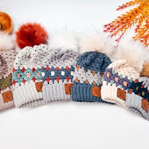May include: A collection of six crocheted beanies featuring granny square patterns in various color combinations, including beige, grey, blue, and cream. Each hat is topped with a faux fur pom-pom and includes a small brown leather tag with the letters RQ.