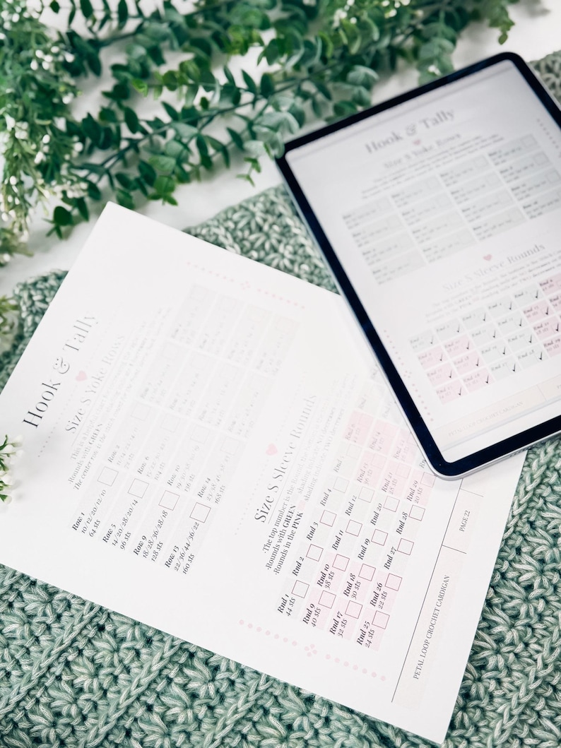 May include: A crochet pattern and a tablet displaying a crochet chart are on a green crocheted blanket. The pattern is titled "Hook & Tally" and includes size and round details. The tablet shows a similar chart. Green foliage is in the background.