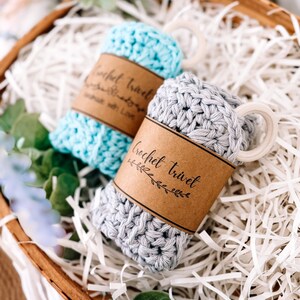 Könnte beinhalten: Zwei gehäkelte Untersetzer, einer hellblau und einer grau, werden in einem Holzkörbchen mit weißem Papier präsentiert. Jeder Untersetzer hat ein braunes Papierband mit dem Text "Crochet trivet" und einen Holzringgriff. Grüne Blätter und Blumen umgeben den Korb.