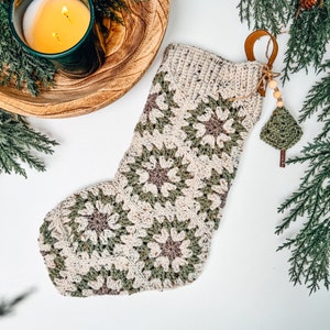 May include: A crochet Christmas stocking constructed from hexagonal motifs in cream, sage green, and muted brown yarn. The stocking features a ribbed cuff and a decorative hanging charm consisting of wooden beads and a small green crochet tree.