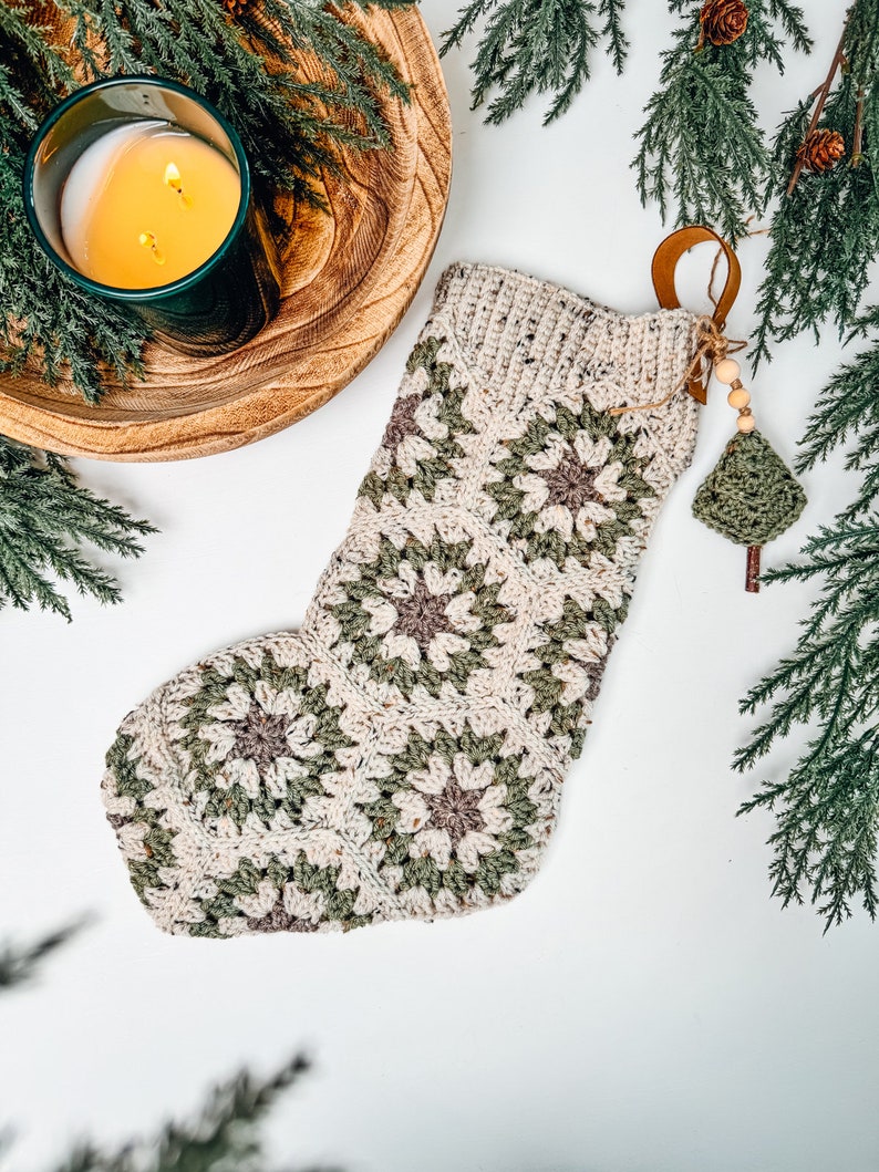 May include: A crochet Christmas stocking constructed from hexagonal motifs in cream, sage green, and muted brown yarn. The stocking features a ribbed cuff and a decorative hanging charm consisting of wooden beads and a small green crochet tree.