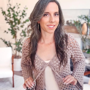 May include: A light brown, long-sleeved crochet cardigan featuring an open-front design with a tie closure at the chest. The garment is crafted with a repeating open-work stitch pattern, long sleeves, and a solid ribbed hem along the bottom edge.