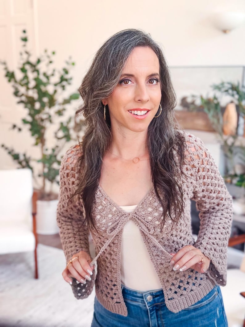 May include: A light brown, long-sleeved crochet cardigan featuring an open-front design with a tie closure at the chest. The garment is crafted with a repeating open-work stitch pattern, long sleeves, and a solid ribbed hem along the bottom edge.
