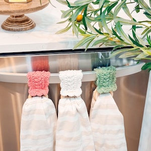 May include: Three kitchen towels with crocheted tops in pink, white, and green hang from a stainless steel appliance handle. The towels have a striped pattern. Olive branches and a wooden cake stand are in the background.