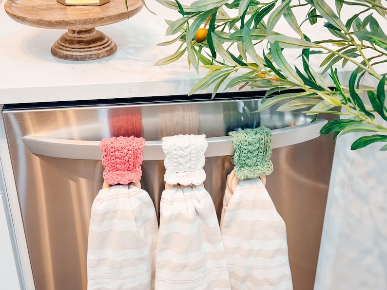 May include: Three kitchen towels with crocheted tops in pink, white, and green hang from a stainless steel appliance handle. The towels have a striped pattern. Olive branches and a wooden cake stand are in the background.