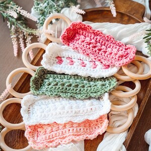 May include: A collection of colorful, crocheted baby toys in pink, white, and green. The toys are rectangular and feature intricate stitch patterns. Wooden rings are arranged around the toys on a wooden tray, with white fabric and greenery in the background.