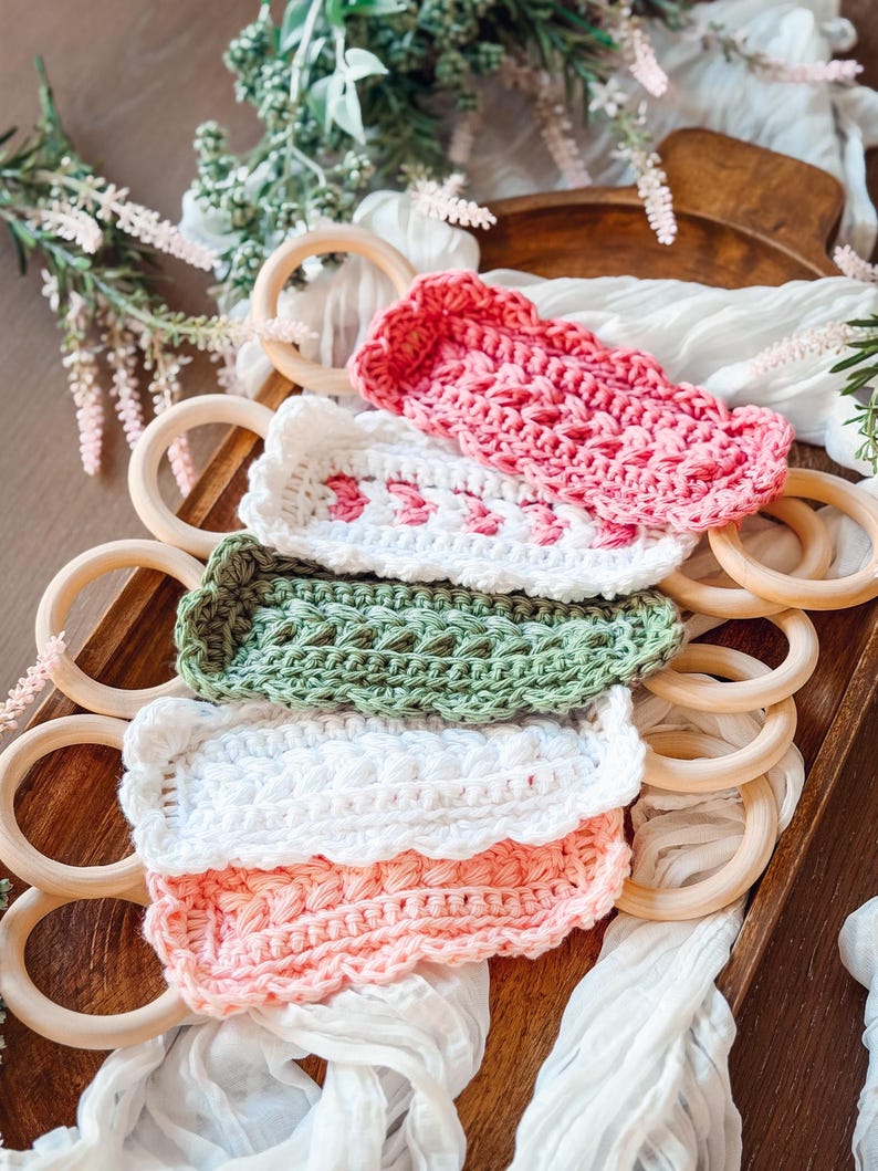 May include: A collection of colorful, crocheted baby toys in pink, white, and green. The toys are rectangular and feature intricate stitch patterns. Wooden rings are arranged around the toys on a wooden tray, with white fabric and greenery in the background.