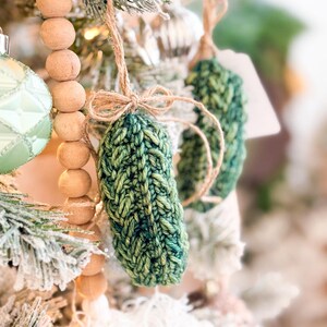 May include: A hand-knitted green pickle ornament featuring a textured yarn stitch pattern. The ornament is suspended by a thin brown twine string tied into a small bow at the top, hanging from a branch alongside other holiday tree decorations.