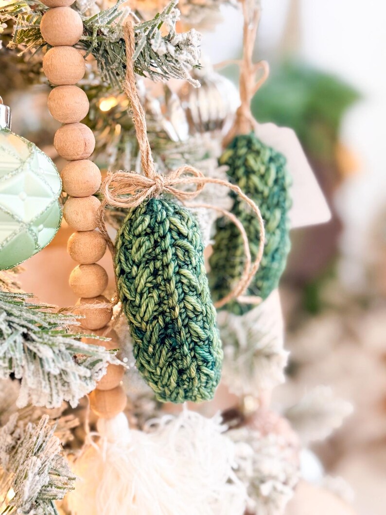 May include: A hand-knitted green pickle ornament featuring a textured yarn stitch pattern. The ornament is suspended by a thin brown twine string tied into a small bow at the top, hanging from a branch alongside other holiday tree decorations.