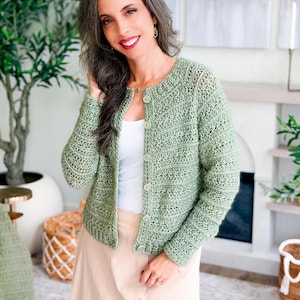 May include: A light green, crocheted cardigan with a round neckline and button closure. The open-weave design of the sweater is visible. The cardigan is worn over a white top and paired with a light-colored skirt. The overall style is casual and handmade.