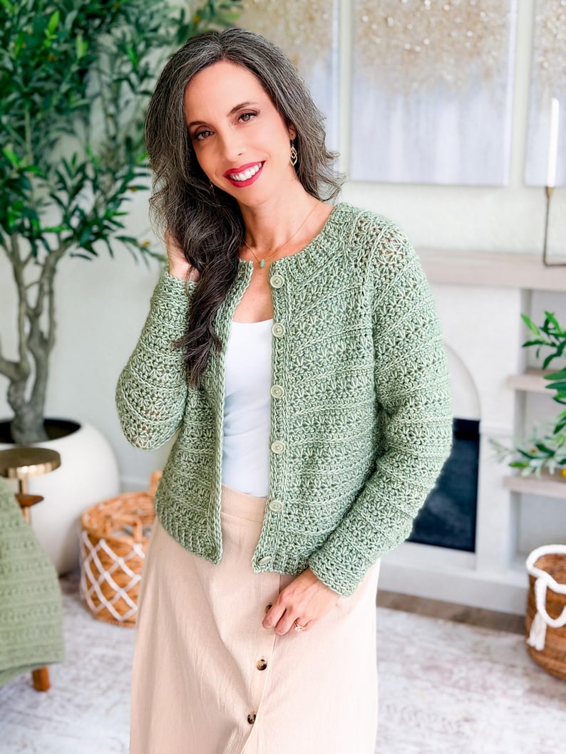 May include: A light green, crocheted cardigan with a round neckline and button closure. The open-weave design of the sweater is visible. The cardigan is worn over a white top and paired with a light-colored skirt. The overall style is casual and handmade.