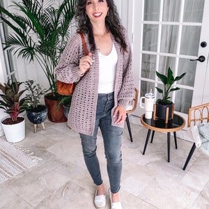 A woman in a white top, grey jeans, and a light crochet cardigan stands smiling in a room with indoor plants and modern decor, showcasing her unique jewellery.