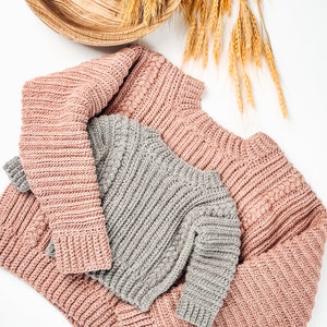 May include: Overhead shot of two knitted sweaters, one dusty rose and one gray, with ribbed and cable knit details. A lit candle in a wooden bowl and wheat stalks are in the background, creating a cozy, autumnal theme.