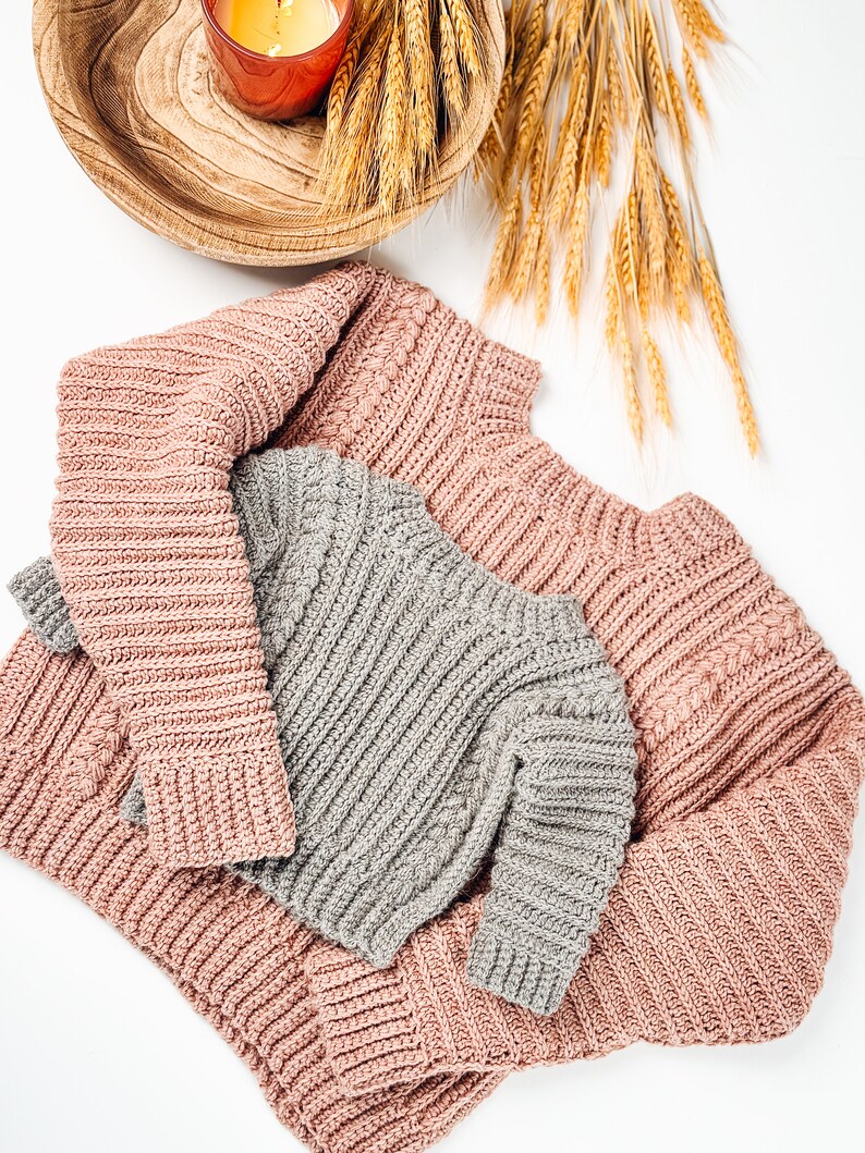 May include: Overhead shot of two knitted sweaters, one dusty rose and one gray, with ribbed and cable knit details. A lit candle in a wooden bowl and wheat stalks are in the background, creating a cozy, autumnal theme.