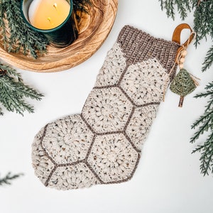 May include: A crochet Christmas stocking constructed from cream-colored hexagonal motifs with brown borders. The top cuff is a ribbed brown knit. A leather hanging loop is attached, adorned with small wooden beads and a miniature green crochet tree.