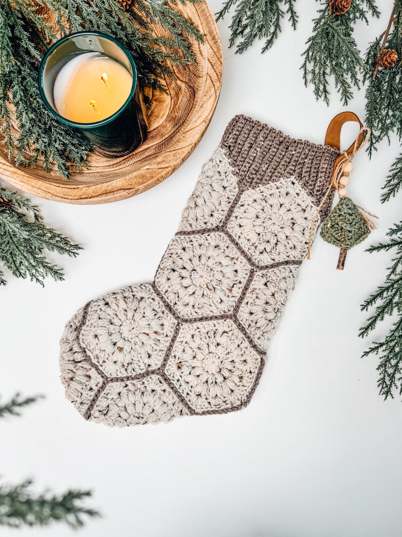 May include: A crochet Christmas stocking constructed from cream-colored hexagonal motifs with brown borders. The top cuff is a ribbed brown knit. A leather hanging loop is attached, adorned with small wooden beads and a miniature green crochet tree.