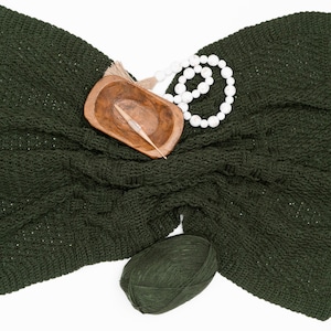 Puede incluir: Una manta de ganchillo en color verde bosque oscuro con un patr&oacute;n texturizado, acompa&ntilde;ada de un peque&ntilde;o cuenco de madera que contiene un ganchillo, un collar de cuentas de madera blanca con borla y un ovillo de lana verde oscuro a juego.