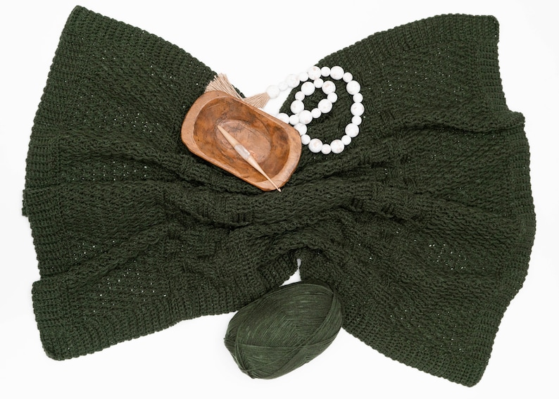 Puede incluir: Una manta de ganchillo en color verde bosque oscuro con un patr&oacute;n texturizado, acompa&ntilde;ada de un peque&ntilde;o cuenco de madera que contiene un ganchillo, un collar de cuentas de madera blanca con borla y un ovillo de lana verde oscuro a juego.