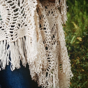 A woman wearing a white shawl decorated with the intricate crochet pineapple stitch, with fringed edges. She stands in an outdoor green space, surrounded by greenery and sunshine.