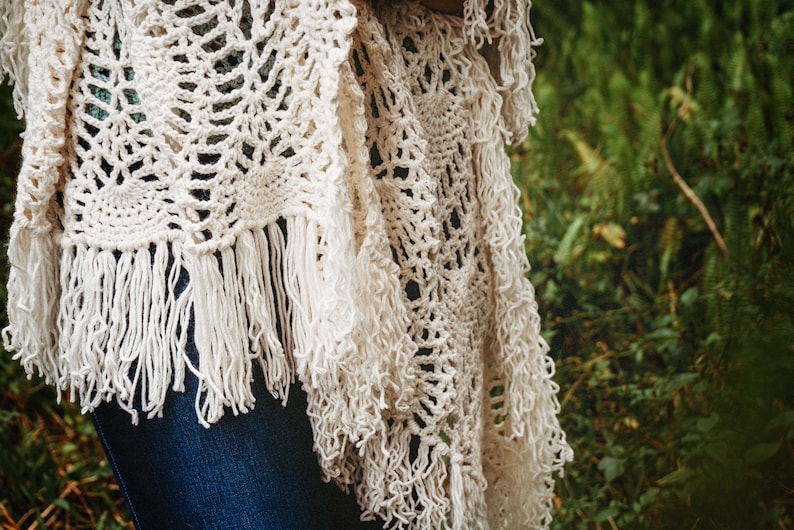 A woman wearing a white shawl decorated with the intricate crochet pineapple stitch, with fringed edges. She stands in an outdoor green space, surrounded by greenery and sunshine.