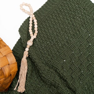 Puede incluir: Una manta de ganchillo de color verde oscuro con un patr&oacute;n texturizado, una cesta de madera tejida con un asa negra y un collar de cuentas de madera clara con una borla de yute beige, todo dispuesto sobre una superficie blanca lisa.