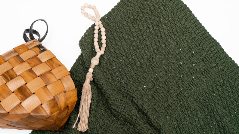 Puede incluir: Una manta de ganchillo de color verde oscuro con un patr&oacute;n texturizado, una cesta de madera tejida con un asa negra y un collar de cuentas de madera clara con una borla de yute beige, todo dispuesto sobre una superficie blanca lisa.