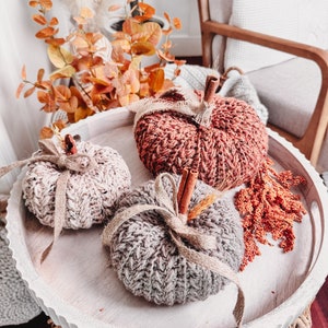 Crocheted pumpkins with cinnamon stick stems are displayed on a round tray, surrounded by autumn foliage, and placed on a woven table—creating the perfect handmade gift.