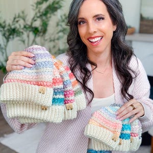 A woman with long dark hair and gray streaks is holding several crochet pastel-colored hats, smiling at the camera indoors, showcasing the perfect handmade gift.