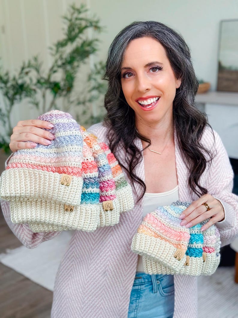 A woman with long dark hair and gray streaks is holding several crochet pastel-colored hats, smiling at the camera indoors, showcasing the perfect handmade gift.